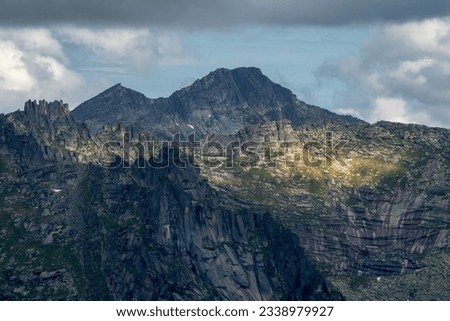 Sunlight glides along the slopes of majestic high and sharp mountains from each side. The peaks are shrouded in rainy haze. Remote and isolated place. Western Sayans. Royalty-Free Stock Photo #2338979927