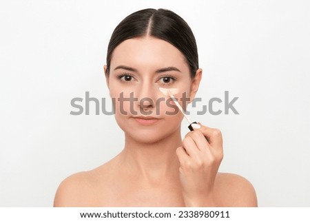 A young attractive Caucasian dark-haired woman applying a concealer under her eyes from dark circles under her eyes. Isolated on a light background Royalty-Free Stock Photo #2338980911