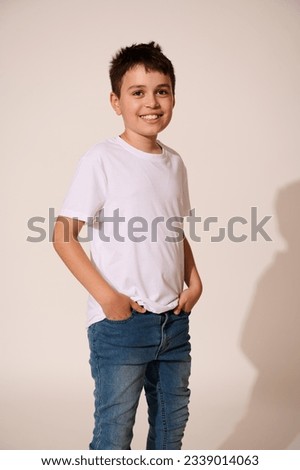 Vertical studio shot of handsome teenager, school boy wearing white mockup t-shirt and blue denim jeans, smiling broadly looking at camera, standing with hands in pockets on white isolated background