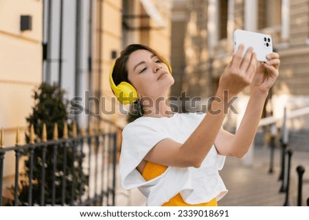 stylish pretty woman wearing yellow headphones listening to music taking pictures on smartphone camera walking in city street, summer style fashion trend