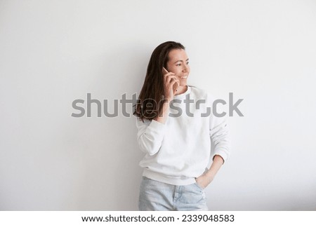 Young woman holding a mobile phone, smiling, using social media. Millennial person in white sweatshirt on white background. People using technology. Work call. 