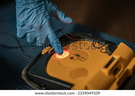First aid training – The instructor is activating the shock. The background is blurred. the focus is on the AED and the shock button. the picture is very detailed. Blue gloves are in the foreground. Royalty-Free Stock Photo #2339049109