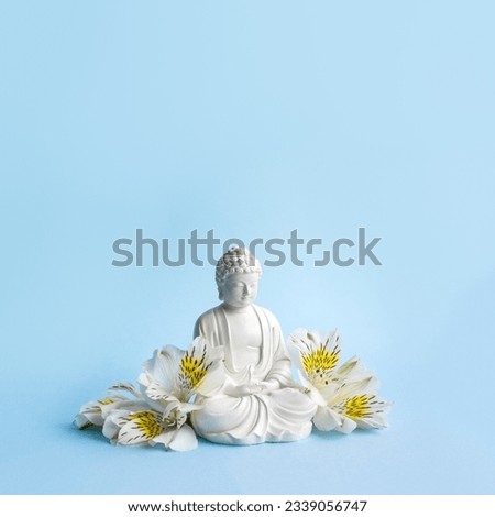 Buddha figurine surrounded by white flowers on blue background. Mental health and meditation concept. Square orientation, selective soft focus.