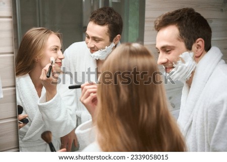 Woman putting on makeup standing in the bathroom man looking at her