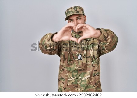 Young hispanic man wearing camouflage army uniform smiling in love doing heart symbol shape with hands. romantic concept. 