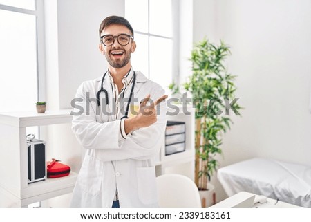 Hispanic man wearing doctor uniform and stethoscope smiling cheerful pointing with hand and finger up to the side 