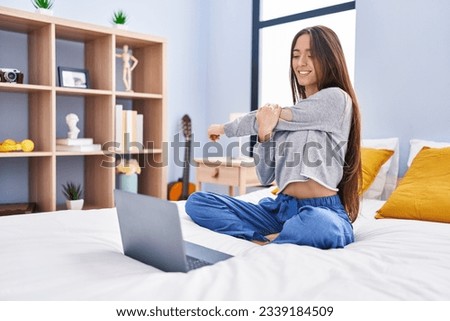Young beautiful hispanic woman doing online stretching exercise sitting on bed at bedroom