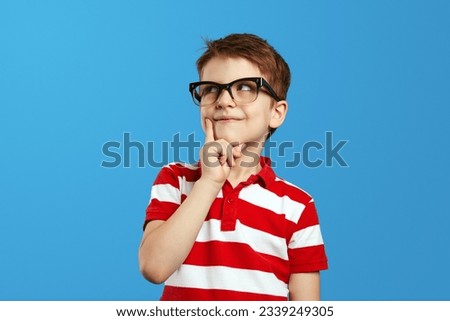 Smart thoughtful preschool boy in nerdy glasses and red striped polo shirt touching cheek and looking up while thinking against blue background.