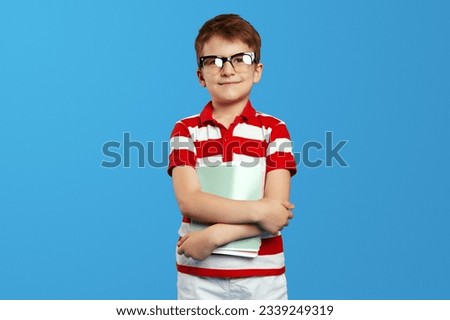Happy little schoolboy in nerdy glasses holding notepads and smiling at camera while standing against blue background, wearing red striped polo shirt.