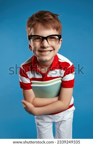 Close up portrait of smart pupil wunderkind in nerdy glasses and red striped shirt looking straight at camera while holding textbooks, standing isolated over blue background. Vertical photo