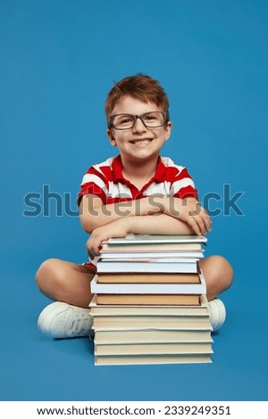 Smart little genius with nerdy eyeglasses holding hands on bunch of books while looking at camera, wearing red striped shirt, isolated over blue background. Back to school concept