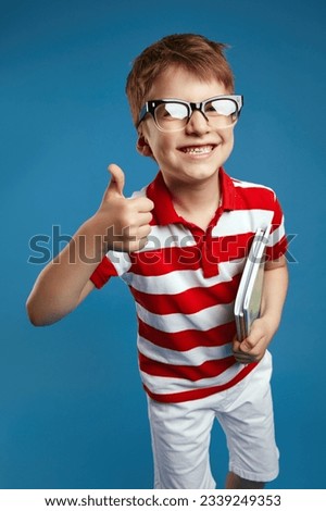 Close up portrait of nerdy boy in retro eyeglasses and red striped shirt holding textbooks while showing thumb up gesture, standing isolated over blue background