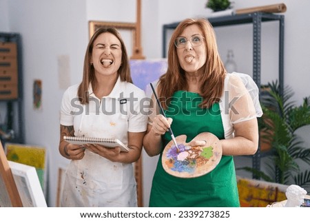 Hispanic mother and daughter painting at paint studio sticking tongue out happy with funny expression. 