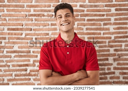 Young hispanic man standing over bricks wall happy face smiling with crossed arms looking at the camera. positive person. 