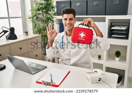 Young hispanic doctor man holding first aid kit doing ok sign with fingers, smiling friendly gesturing excellent symbol 
