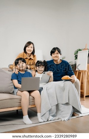 Asian Overjoyed kids sitting on sofa with cheerful parents, watching funny video on computer. Happy married couple enjoying spending weekend time with children, looking at laptop and tablet  Royalty-Free Stock Photo #2339280989