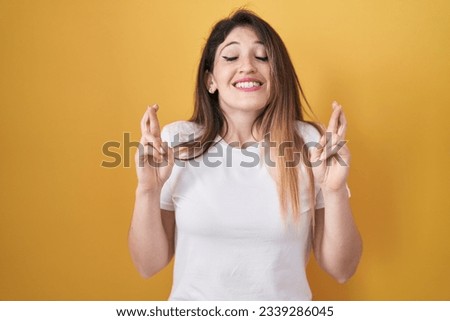 Young brunette woman standing over yellow background gesturing finger crossed smiling with hope and eyes closed. luck and superstitious concept. 