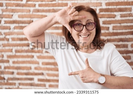 Senior woman with glasses standing over bricks wall smiling making frame with hands and fingers with happy face. creativity and photography concept. 