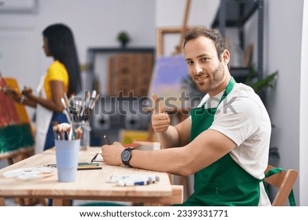 Hispanic man at painter studio smiling happy and positive, thumb up doing excellent and approval sign 