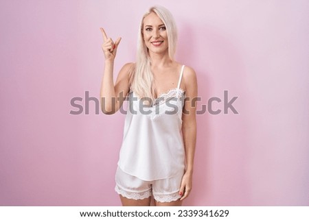 Caucasian woman wearing pajama wearing pink background cheerful with a smile on face pointing with hand and finger up to the side with happy and natural expression 