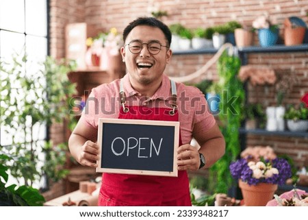 Chinese young man working at florist holding open sign smiling and laughing hard out loud because funny crazy joke. 