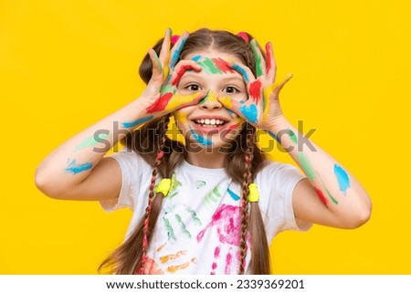 A little girl stained with multicolored paints. A happy, beautiful young girl painted with artistic paints. The child makes glasses out of his fingers and smiles. Yellow isolated background.