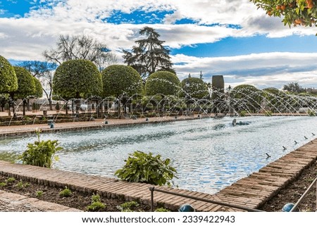 Square fountain with jets coming out of spouts in gardens of Alcazar de los Reyes Cristianos, small trees pruned in circular shape in background, day with blue sky cloud-capped in Cordoba, Spain