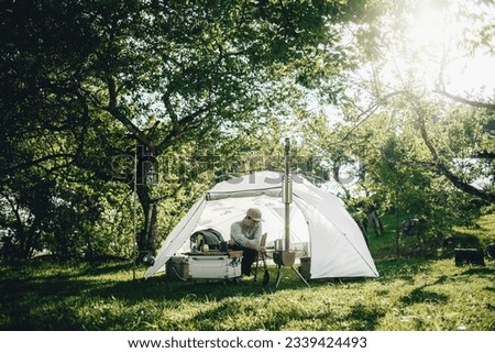 A man with a white glamping tent in the forest with trees and green grass, with camping chair, table and equipments in the tent. there's sunlight and clear sky with golden hour vibes in the morning