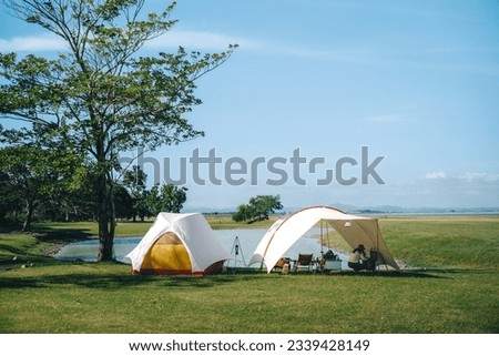 White tent and tarp next to a pond with green grass and trees and blue sky with clouds, it's golden hour in the morning vibes in luxury camping or glamping Royalty-Free Stock Photo #2339428149