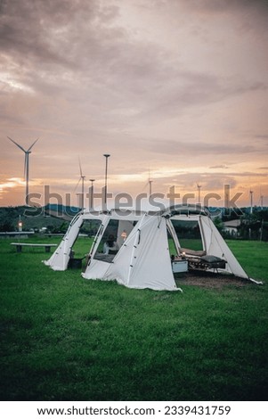 A white tent on green grass during twilight time in the evening with windmills and mountains and camping gears. The sun is about to set