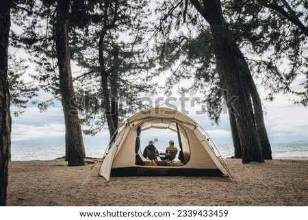 A man and a woman sitting in a white tent among trees and sand with a view of the sea during daytime with sunlight and blue sky Royalty-Free Stock Photo #2339433459