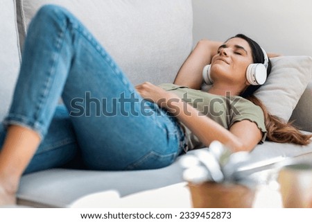 Shot of beautiful kind woman relaxing while listening music with headphones lying on couch at home Royalty-Free Stock Photo #2339452873