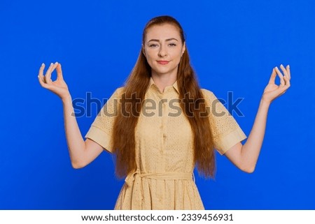 Keep calm down, relax, inner balance. Young redhead woman breathes deeply with mudra gesture, eyes closed, meditating with concentrated thoughts, peaceful mind. Adult girl isolated on blue background