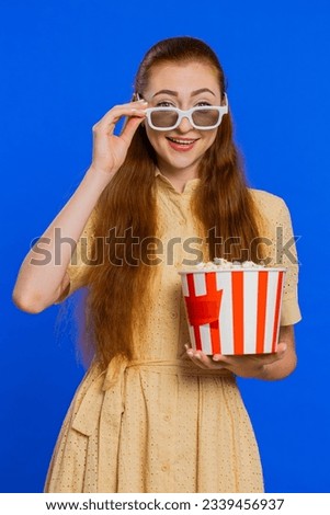 Excited young adult woman in 3D glasses eating popcorn and watching interesting tv serial, sport game film, online social media movie content. Redhead girl isolated on blue studio background. Vertical