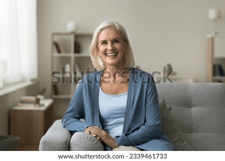 Happy pretty senior woman sitting on couch at cozy home, looking at camera with toothy smile. Positive blonde older lady enjoying leisure on comfortable sofa, posing for indoor portrait Royalty-Free Stock Photo #2339461733