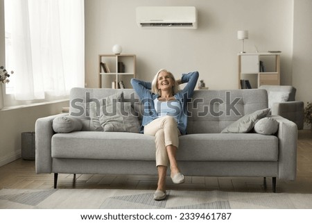 Cheerful relaxed retired old woman resting on grey soft couch under domestic conditioner in stylish cozy apartment interior, breathing cool fresh air, cold comfortable temperature in living room Royalty-Free Stock Photo #2339461787