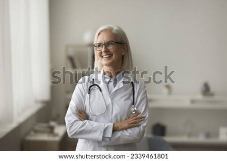 Positive older mature practitioner woman in glasses posing indoors, looking at window away, thinking, smiling. Senior medical professional, doctor in white uniform casual portrait