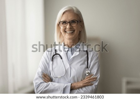 Happy confident senior doctor woman in glasses and white uniform coat posing with crossed hands, looking at camera, smiling. Mature practitioner, medical worker professional portrait