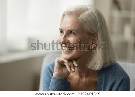 Cheerful positive blonde senior woman sitting on sofa, thinking on good retirement plans, touching chin, looking away, smiling, laughing. Happy elderly lady enjoying relaxation, leisure at home Royalty-Free Stock Photo #2339461815