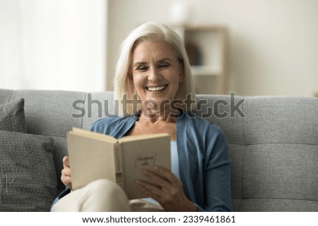 Happy blonde senior retired woman reading paper book, relaxing on home couch, enjoying leisure, relaxation, literature, bestseller novel story, smiling with perfect white teeth, laughing