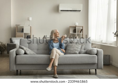 Positive relaxed senior lady using remote control for turning AC on, starting cooling domestic equipment, relaxing on comfortable home couch in cozy modern apartment interior Royalty-Free Stock Photo #2339461865