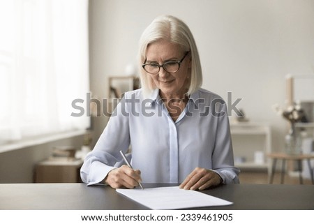 Focused successful mature old business woman in glasses signing contract, agreement at home workplace, writing in papers, reviewing legal documents, sitting at work table
