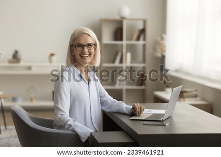 Positive confident senior freelance business woman in glasses posing at home work table, sitting at laptop, looking at camera, smiling, laughing. Retired freelancer, teacher, entrepreneur portrait