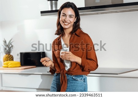 Shot of beautiful happy woman eating a yoghurt while looking at camera in the kitchen at home Royalty-Free Stock Photo #2339463511