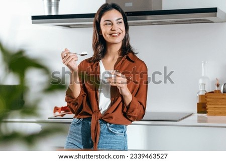 Shot of beautiful happy woman eating a yoghurt while looking forwards in the kitchen at home Royalty-Free Stock Photo #2339463527