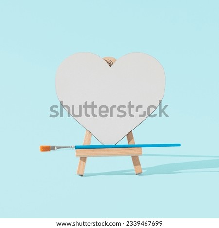 White heart on a wooden painting easel with a brush. Light blue background. Front view. Copy space. Minimal concept of love message.