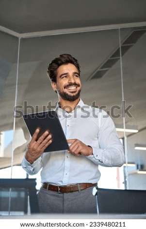 Smiling busy young Latin business man employee using tablet standing in office at work. Happy male professional executive manager using tab computer looking away managing financial data. Vertical Royalty-Free Stock Photo #2339480211
