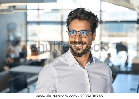 Smiling young Latin business man standing in office looking away, portrait. Happy Hispanic male manager, rich entrepreneur, professional ceo executive wearing eyeglasses thinking of financial success. Royalty-Free Stock Photo #2339480249