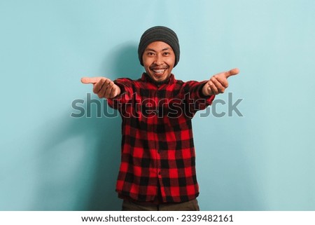 Smiling young Asian man wearing a beanie hat and a red plaid flannel shirt is beckoning with both hands, offering a hug with a come here gesture, inviting, welcoming, isolated on blue background