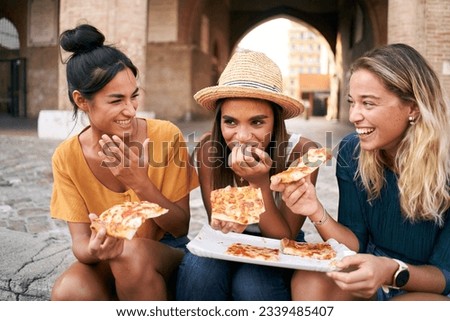 Three beautiful women sitting on the stairs of the city streets eating pizza from a street stall. The happy girls enjoy the weekend together. High quality photo Royalty-Free Stock Photo #2339485407